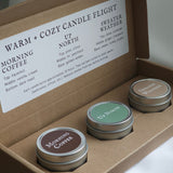 Three small candles in metal tins with labels in a cardboard box, featuring scents like 'Morning Coffee' and 'Sweater Weather'.