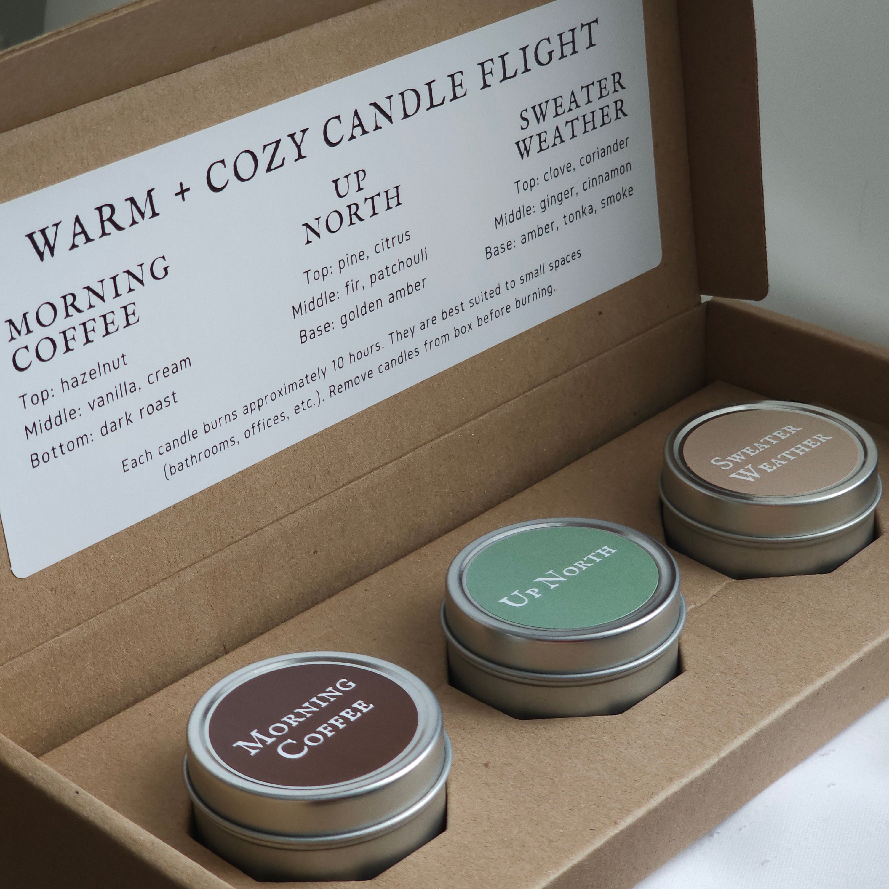 Three small candles in metal tins with labels in a cardboard box, featuring scents like 'Morning Coffee' and 'Sweater Weather'.