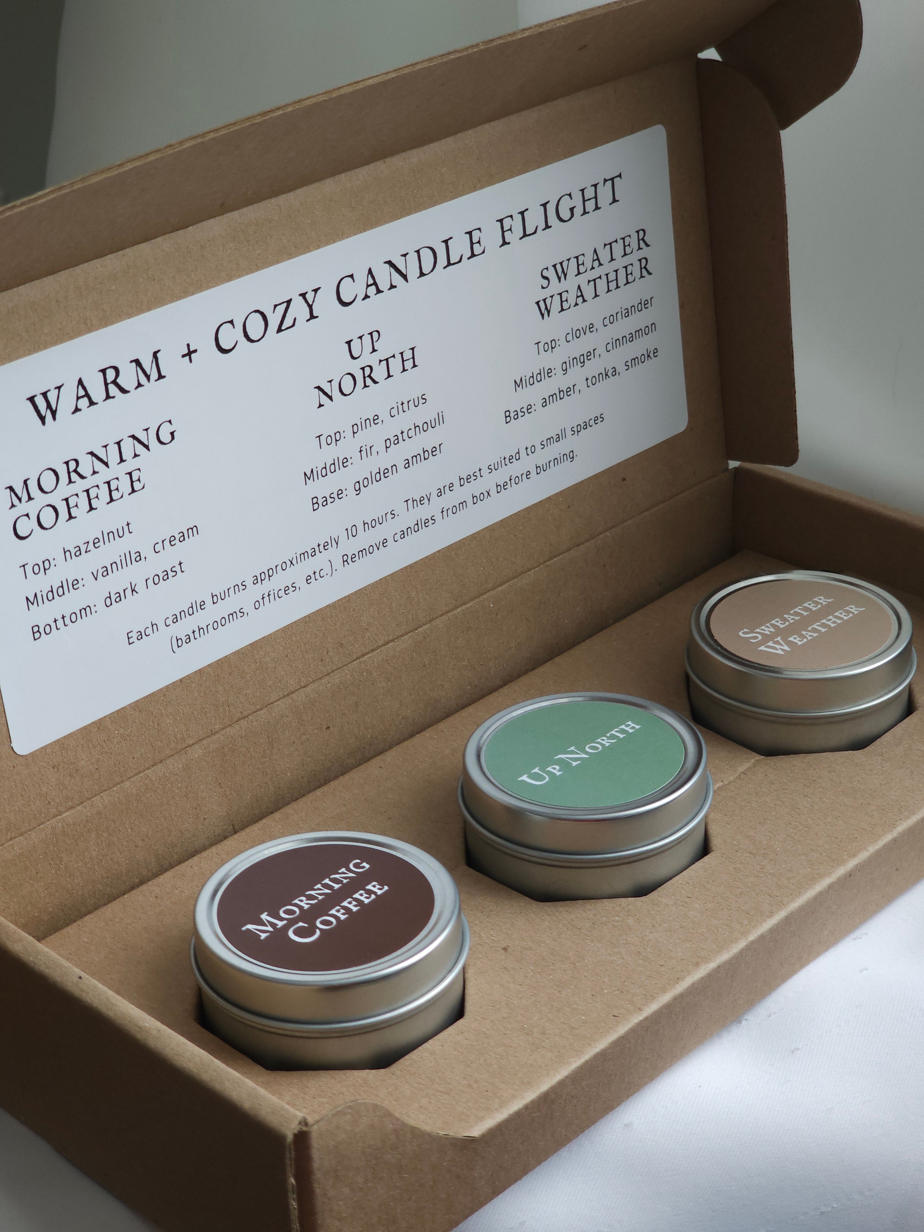 Three small candles in metal tins with labels in a cardboard box, featuring scents like 'Morning Coffee' and 'Sweater Weather'.
