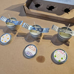 Small candles with lids on a brown surface, with a cardboard box in the background.