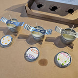 Small candles with lids on a brown surface, with a cardboard box in the background.