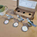 Set of small candles in metal containers with spoons on a cardboard surface, with a 'Handmade Candle Flight' box in the background.