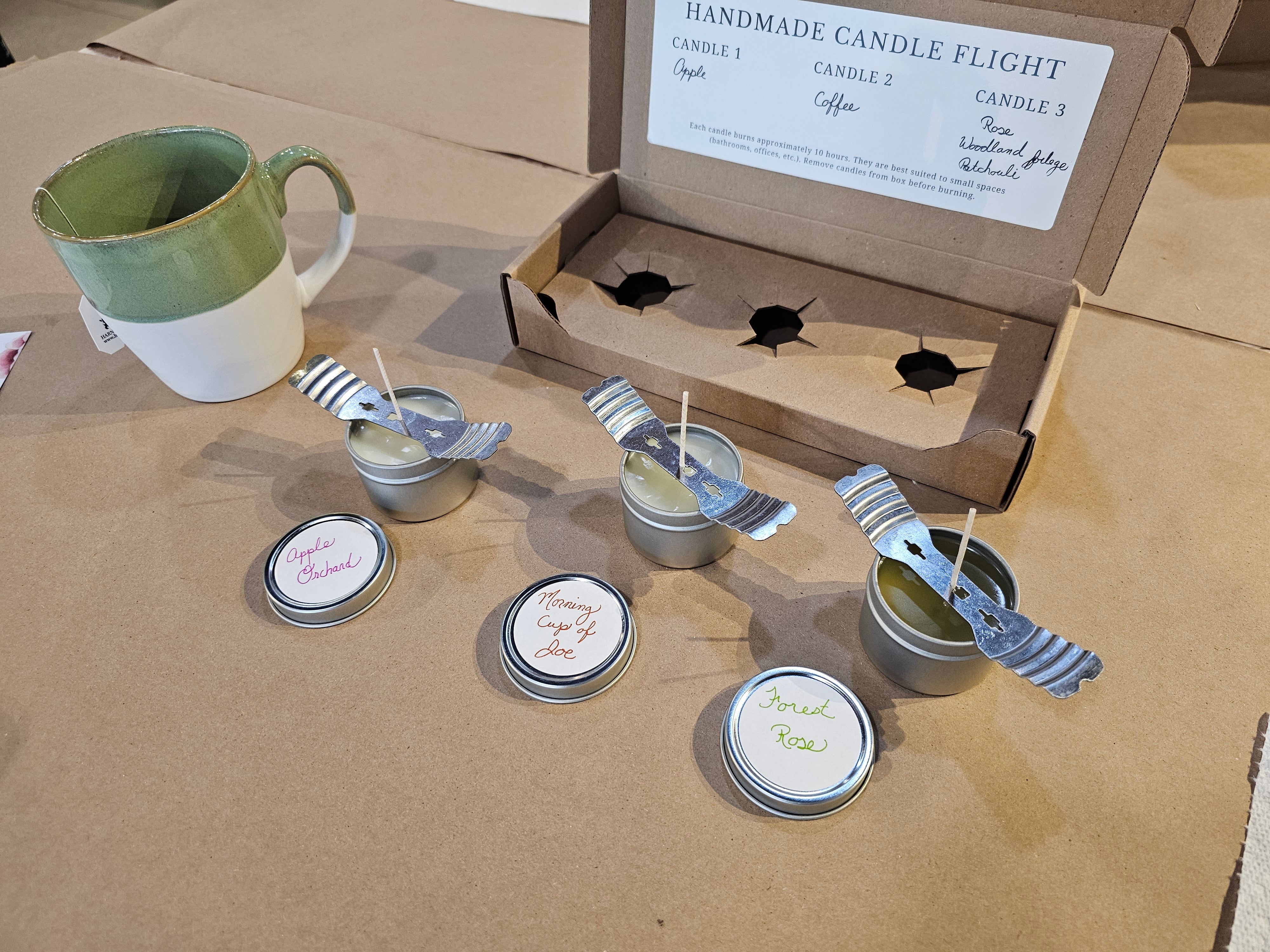 Set of small candles in metal containers with spoons on a cardboard surface, with a 'Handmade Candle Flight' box in the background.